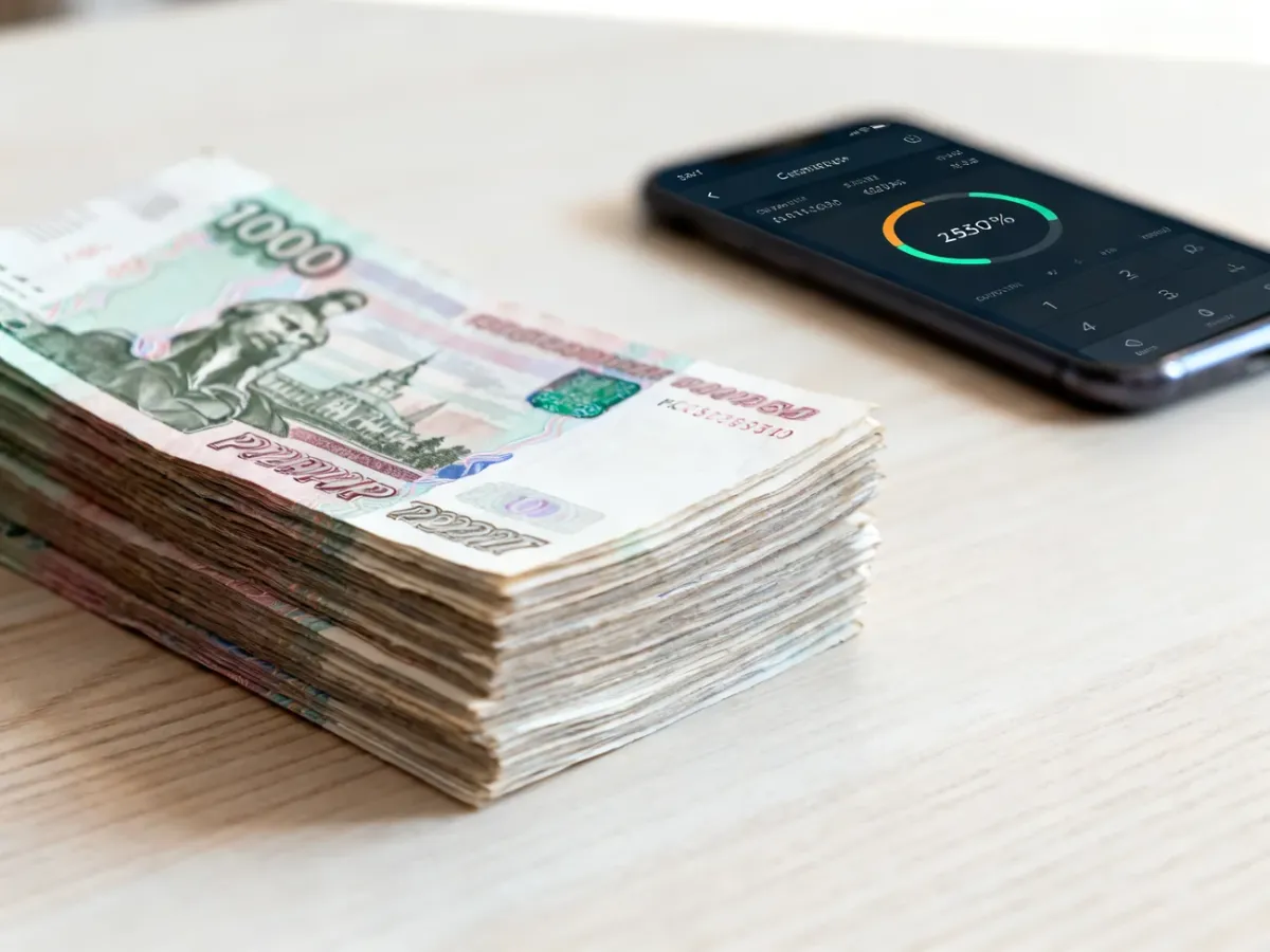 Stos banknotów rubla rosyjskiego na drewnianym stole w tle z rozmytą aplikacją do przeliczania walut na smartfonie.